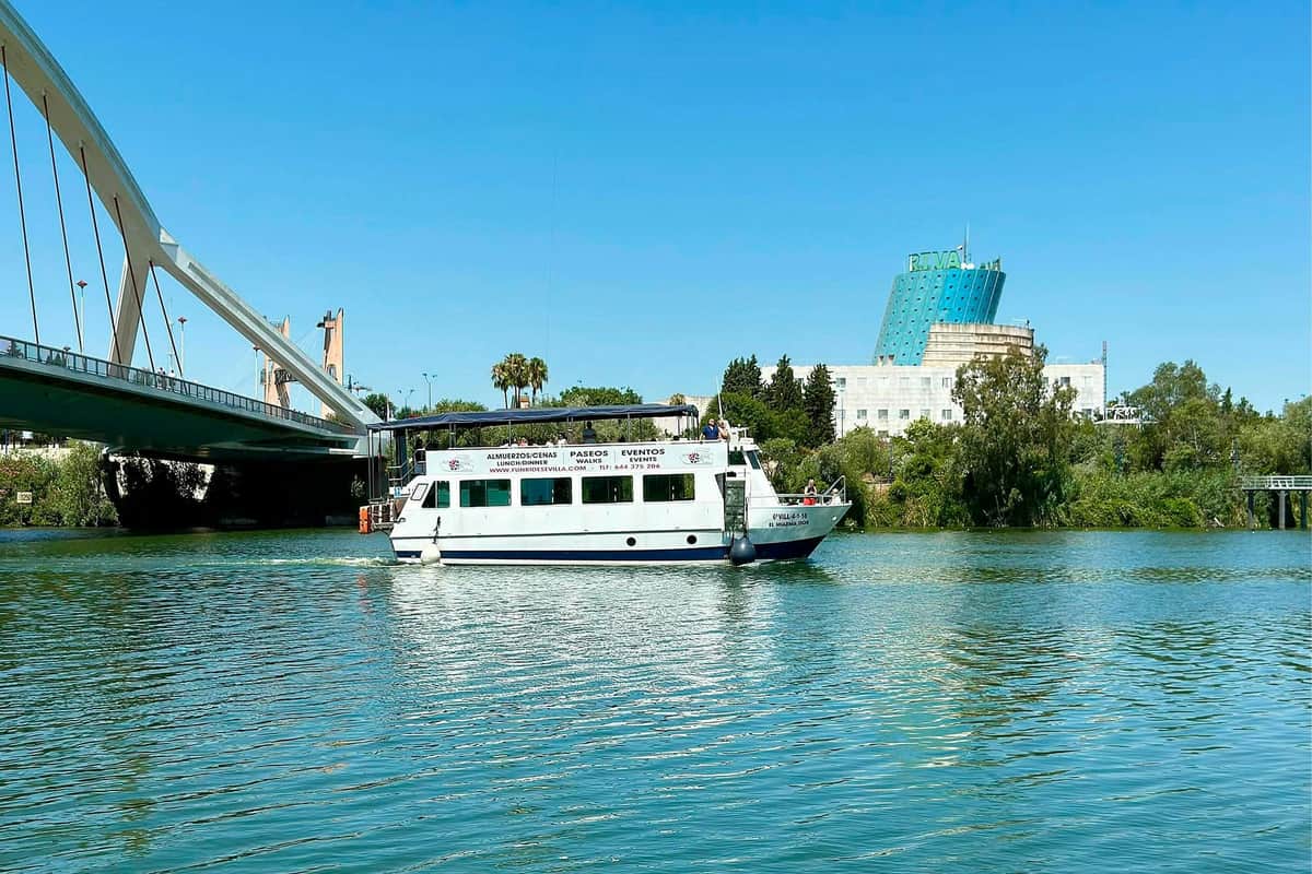 Seville: Guided Guadalquivir River Cruise A white passenger boat on a river with a bridge, trees, and buildings in the background under a clear blue sky.