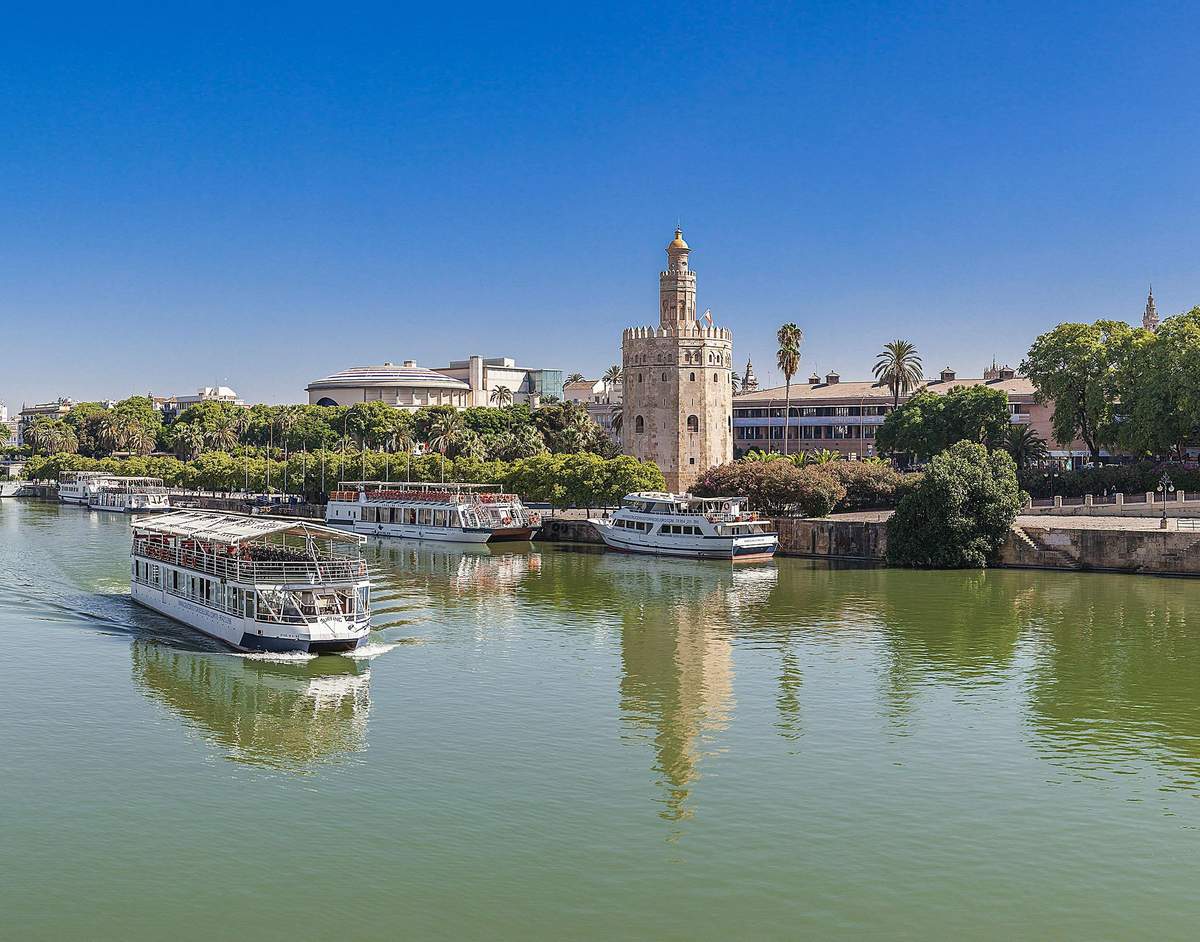 Seville: Sightseeing Cruise from Torre Del Oro Several tour boats on a river with a historic stone tower and buildings surrounded by trees under a clear blue sky.