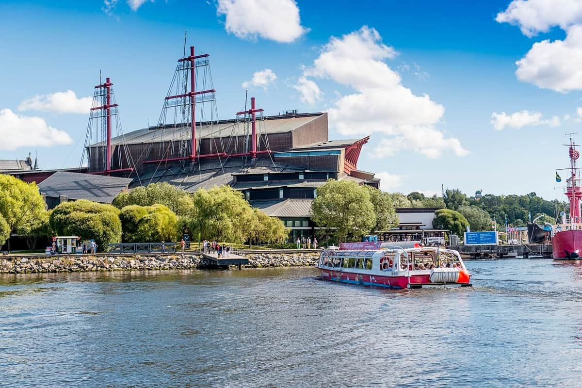Stockholm : visite en bateau Hop-on Hop-off Un bateau d'excursion rouge et blanc sur une rivière, avec un grand bâtiment et des arbres luxuriants le long de la rive, sous un ciel bleu partiellement nuageux.
