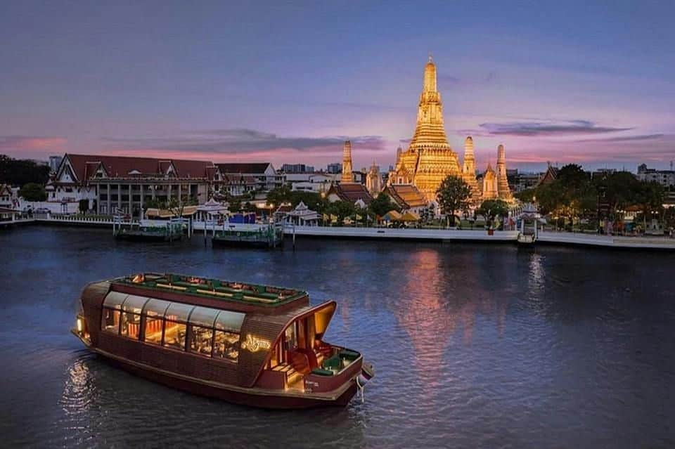 Een boot op een rivier bij zonsondergang, met op de achtergrond een verlichte tempel, omringd door andere gebouwen en bomen.