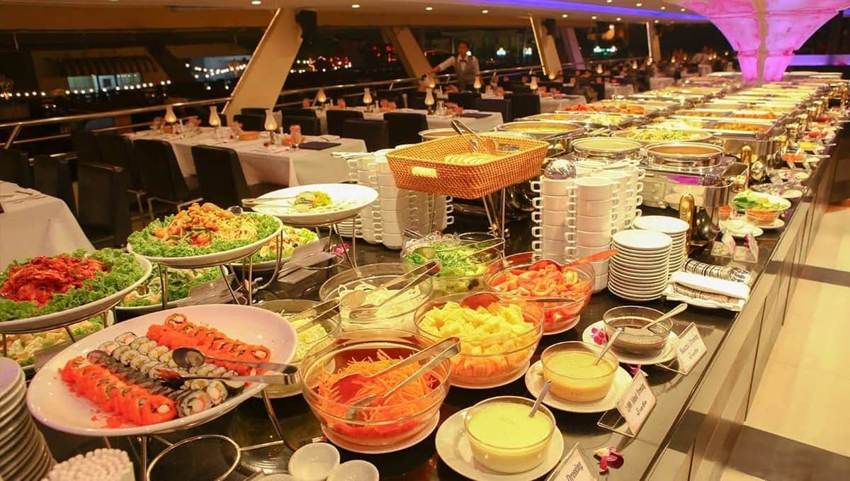 Bangkok: Chao Phraya Princess Dinner Cruise A buffet spread with various salads, fruits, and sauces in bowls. Dining tables and chairs are set up in the background.