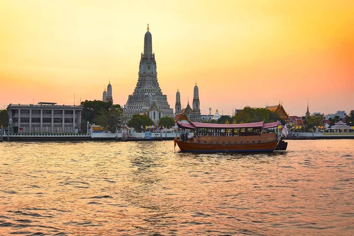 Bangkok: Chao Phraya River Manohra Sunset Dinner Cruise A traditional boat on a river at sunset with a large temple and smaller spires in the background.