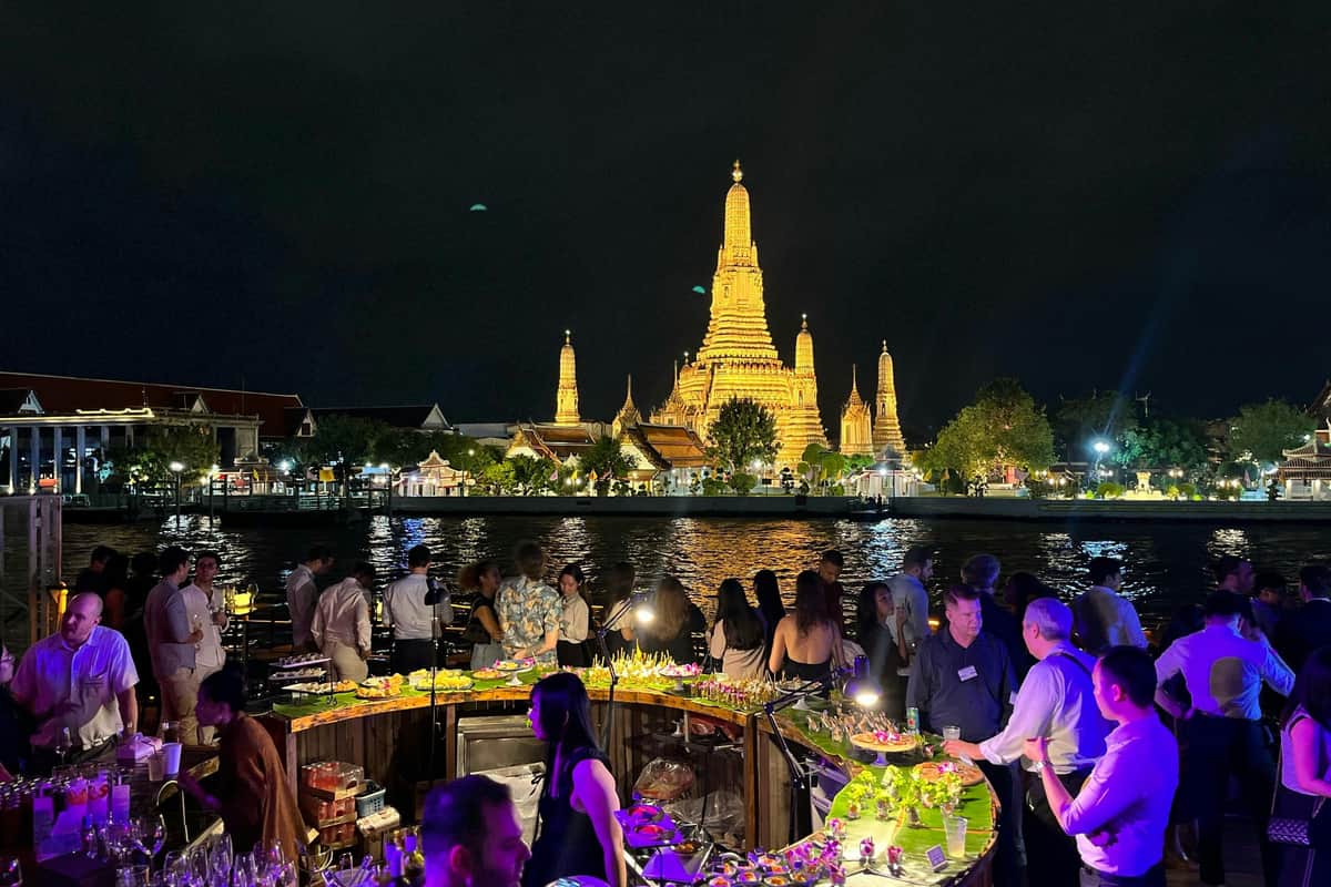 Bangkok: Evening Boat Party + Live DJ + Drink People socializing at an outdoor evening event near a river, with a brightly lit temple in the background.