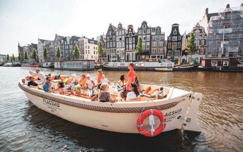 Een groep mensen geniet van een zonnige dag op een rondvaartboot met klassieke Amsterdamse huizen op de achtergrond.