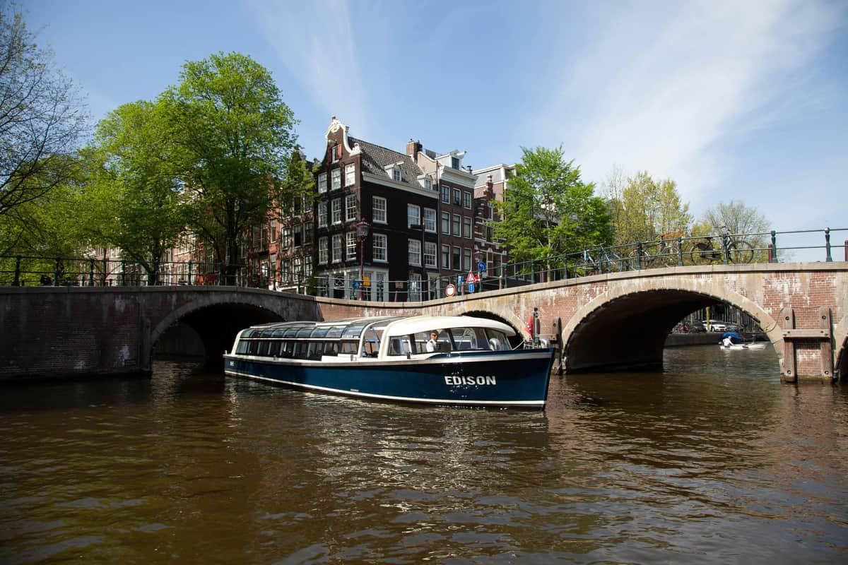Amsterdam: Canal Cruise & Heineken Experience A boat named Edison floating on a canal in front of a bridge and traditional Dutch-style buildings under a clear sky.