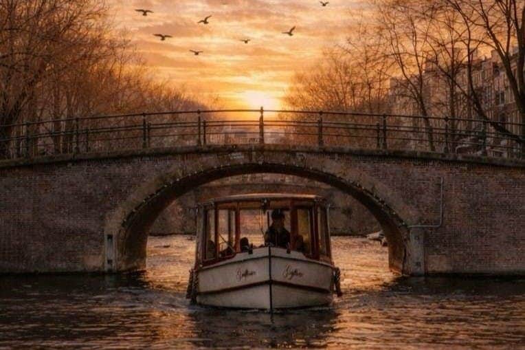 Amsterdam: Abendliche Bootstour auf einem offenen Boot A boat navigating a canal under a stone bridge at sunset, with birds flying in the sky and trees lining the water.