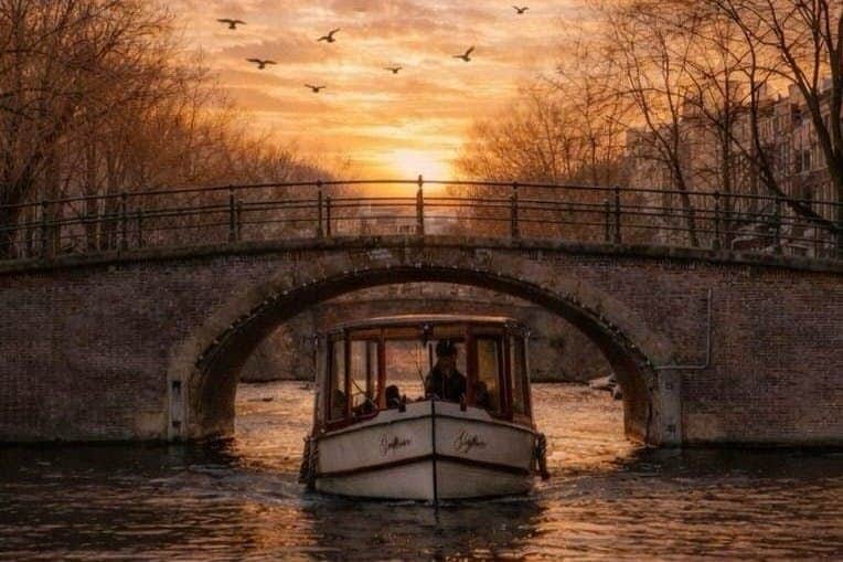 Amsterdam: Flagship Evening Open Boat Cruise A boat passing under a stone bridge on a canal at sunset, with birds flying in the sky and trees lining the canal.