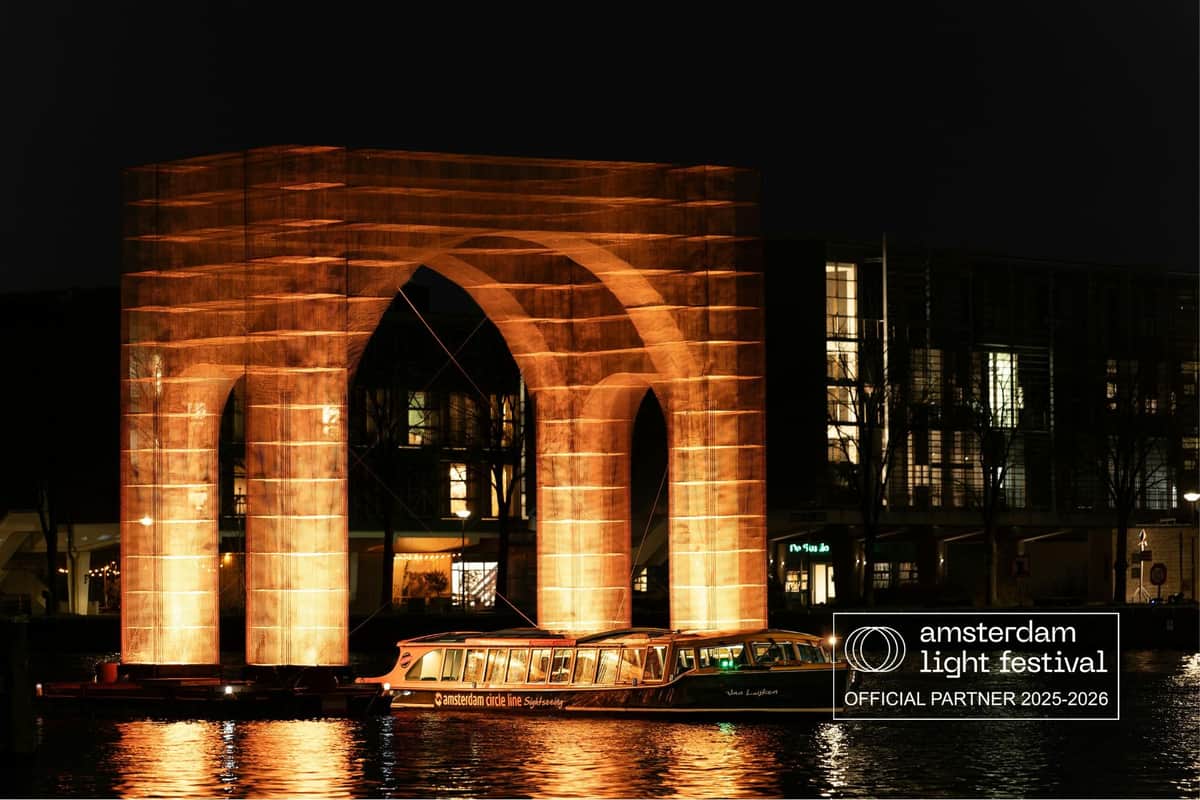 ボートが下を通過し、建物を背景にした、夜の水上のライトアップされたアーチ彫刻。 バナー：「アムステルダム・ライトフェスティバル」。