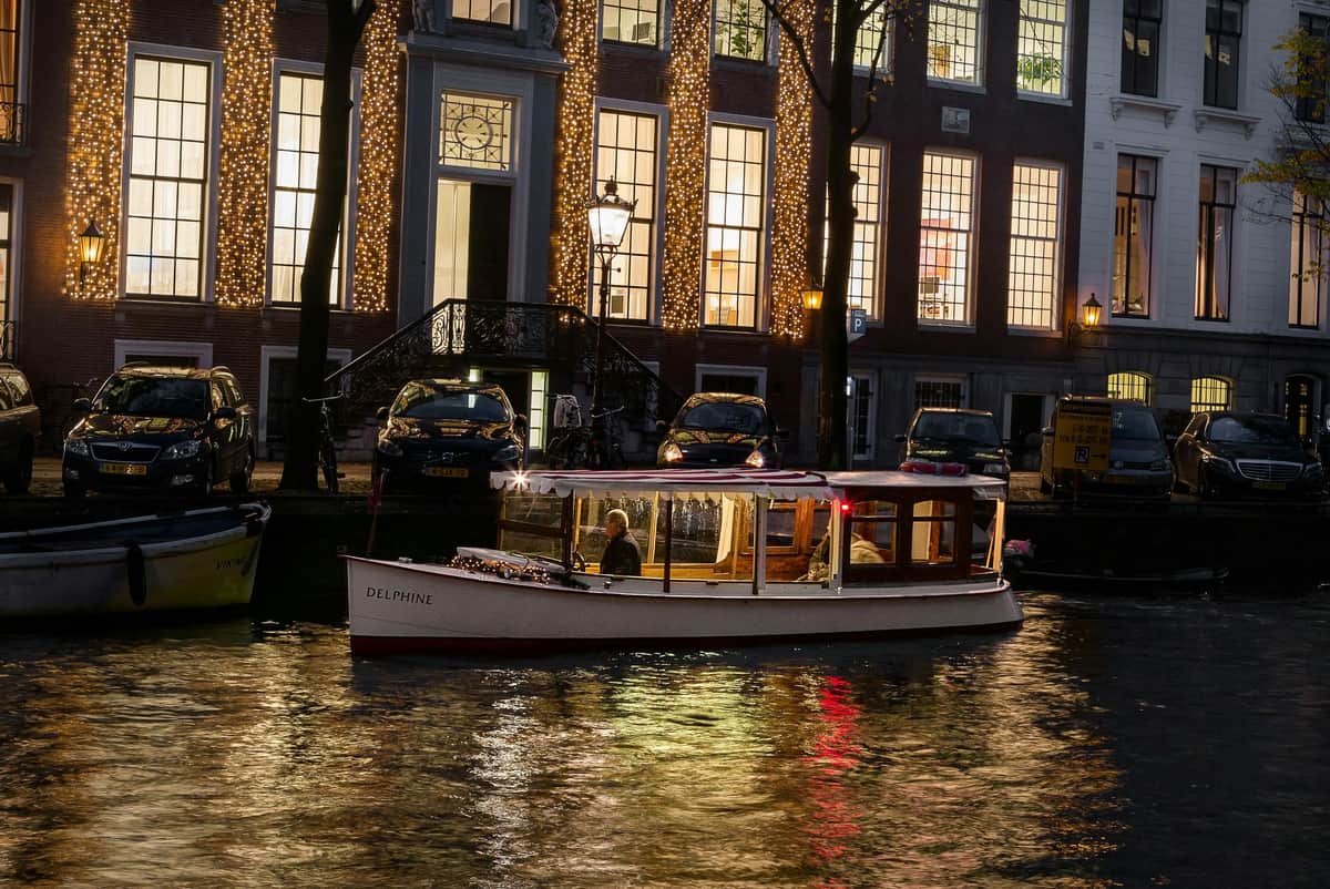 Een boot met de naam "Delphine" op een kanaal met verlichte gebouwen erachter en geparkeerde auto's langs de waterkant in de schemering.