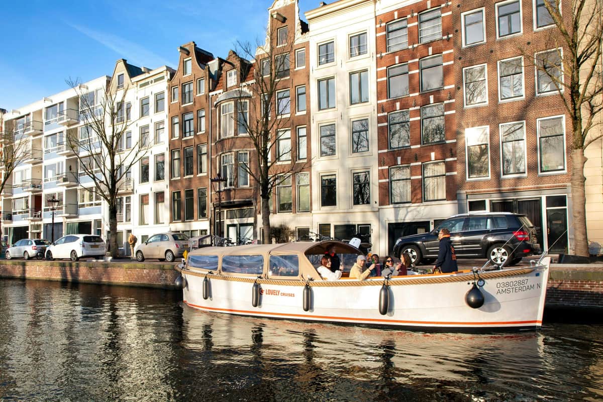 A boat with passengers cruises along a canal in front of classic European townhouses under a clear blue sky.