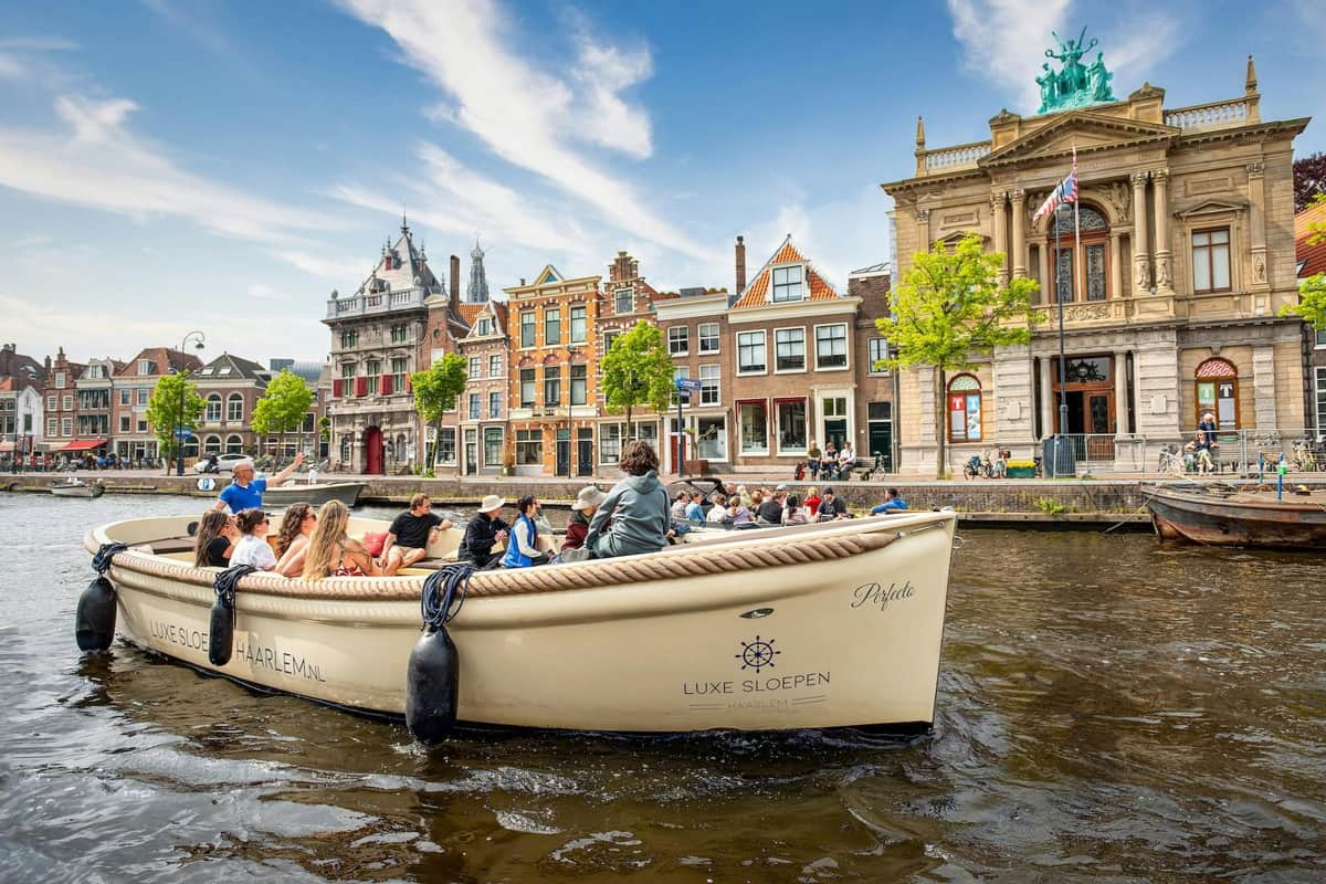 Haarlem : Croisière guidée sur les canaux Un groupe de personnes sur un bateau dans un canal avec des bâtiments historiques et des arbres en arrière-plan sous un ciel partiellement nuageux.