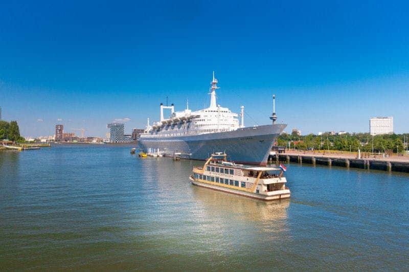 Pancake Boat Rotterdam A large white cruise ship docked at a pier, with a smaller boat cruising nearby, set against a clear blue sky.