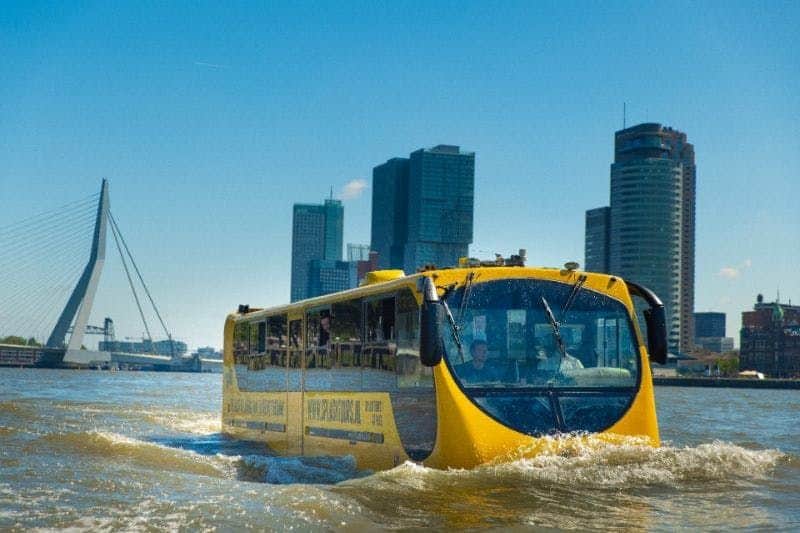Splashtour Rotterdam A yellow amphibious bus travels on water in front of a cityscape with modern skyscrapers under a clear blue sky.