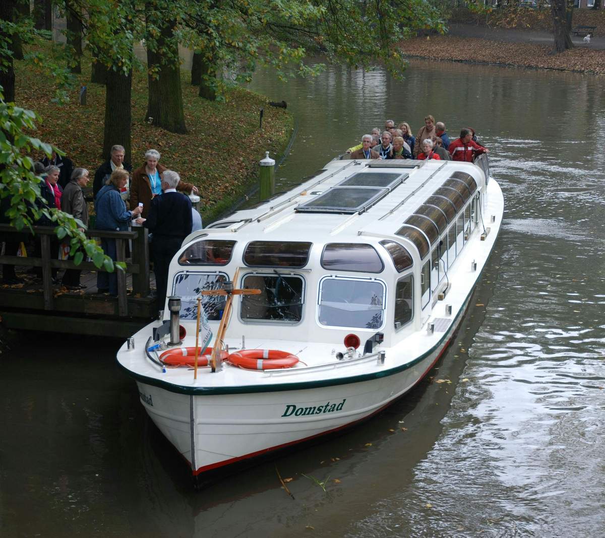 Croisière sur les canaux d'Utrecht Un bateau d'excursion blanc nommé "Domstad" a accosté, avec des passagers à bord et un groupe de personnes debout sur une plate-forme au bord de la rivière.