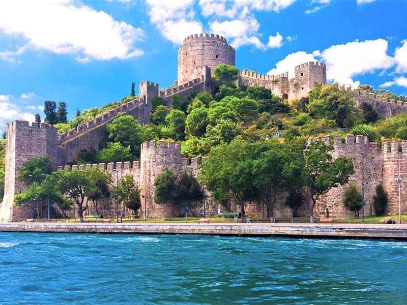 Bosphore, Mer Noire et Palais de Dolmabahce : Excursion en bateau avec déjeuner + entrée Une forteresse historique en pierre avec des tours et des murs sur une colline, entourée de verdure, à proximité d'un plan d'eau.