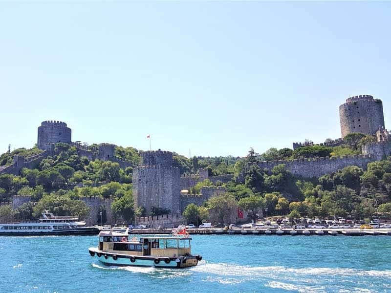 Istanbul: Bosphorus & Black Sea Sightseeing Cruise, Guided Tour + Transport A small boat sails on a blue river in front of a historic fortress on a green hillside under a clear sky.