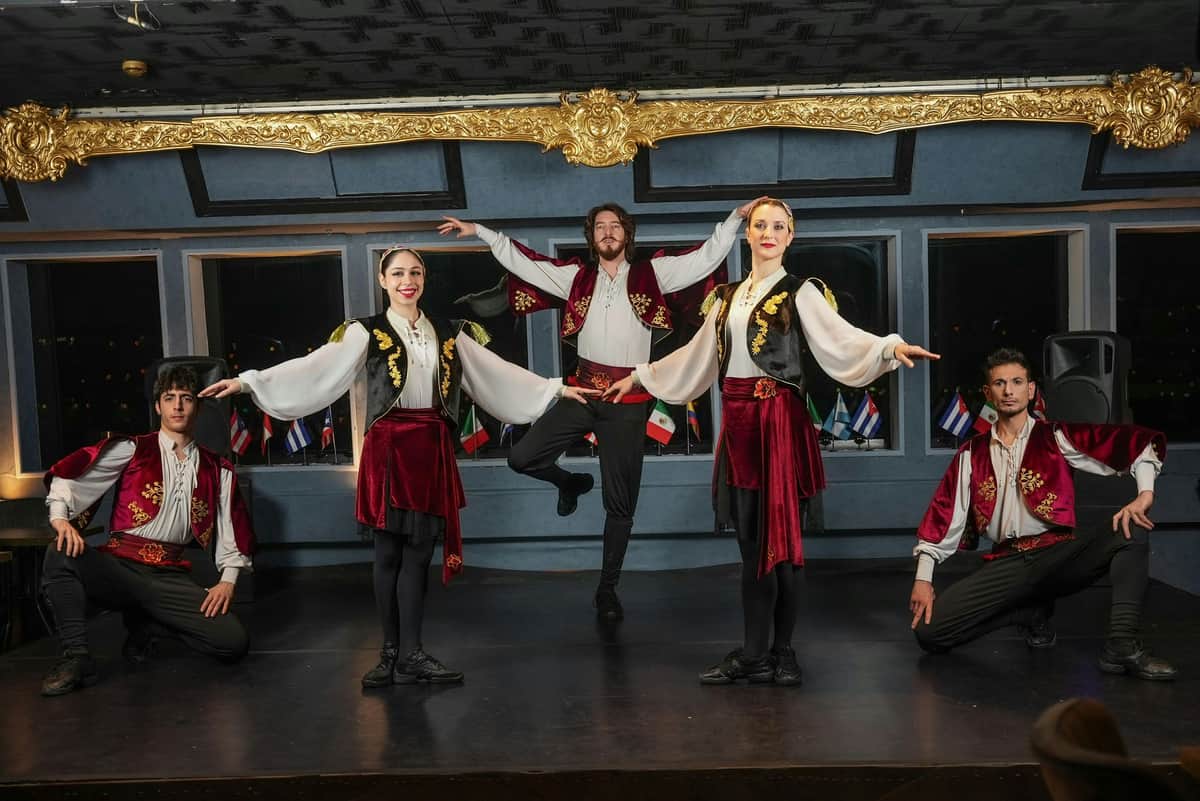 Istanbul: Bosphorus Dinner Cruise with Turkish Dance Shows + Private Table Four dancers in traditional costumes perform on a dimly lit stage with gold trim and flags in the background.