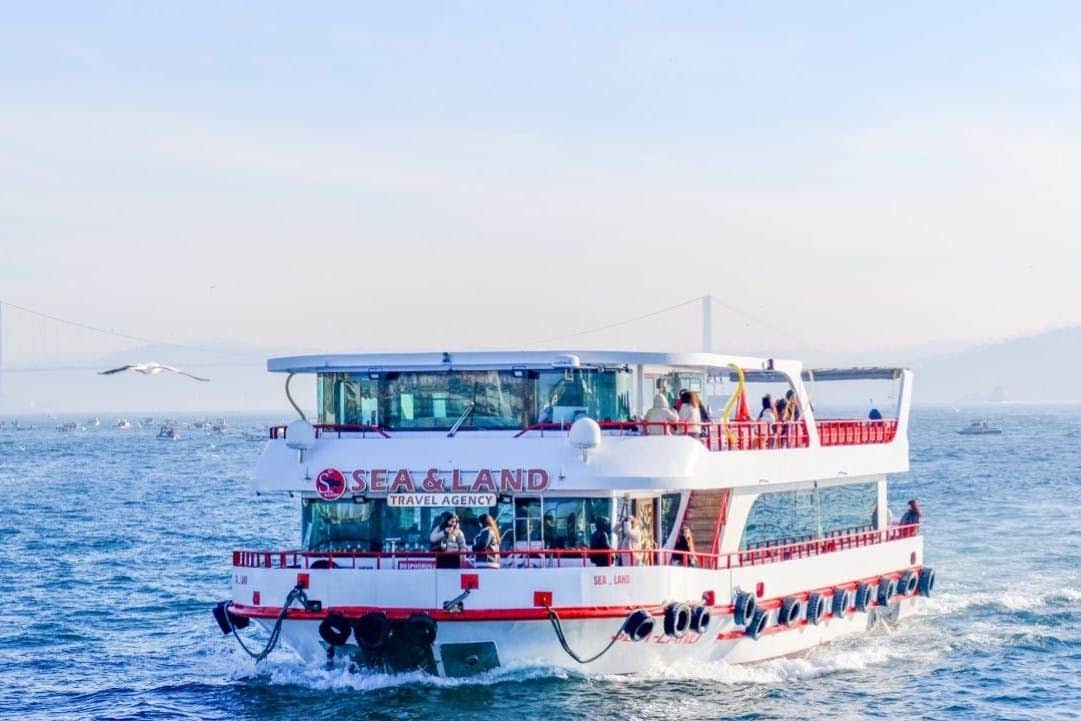 Istanbul: Bosphorus Sightseeing Cruise A white and red cruise boat on a body of water, with people on board enjoying the ride and a bridge visible in the background.