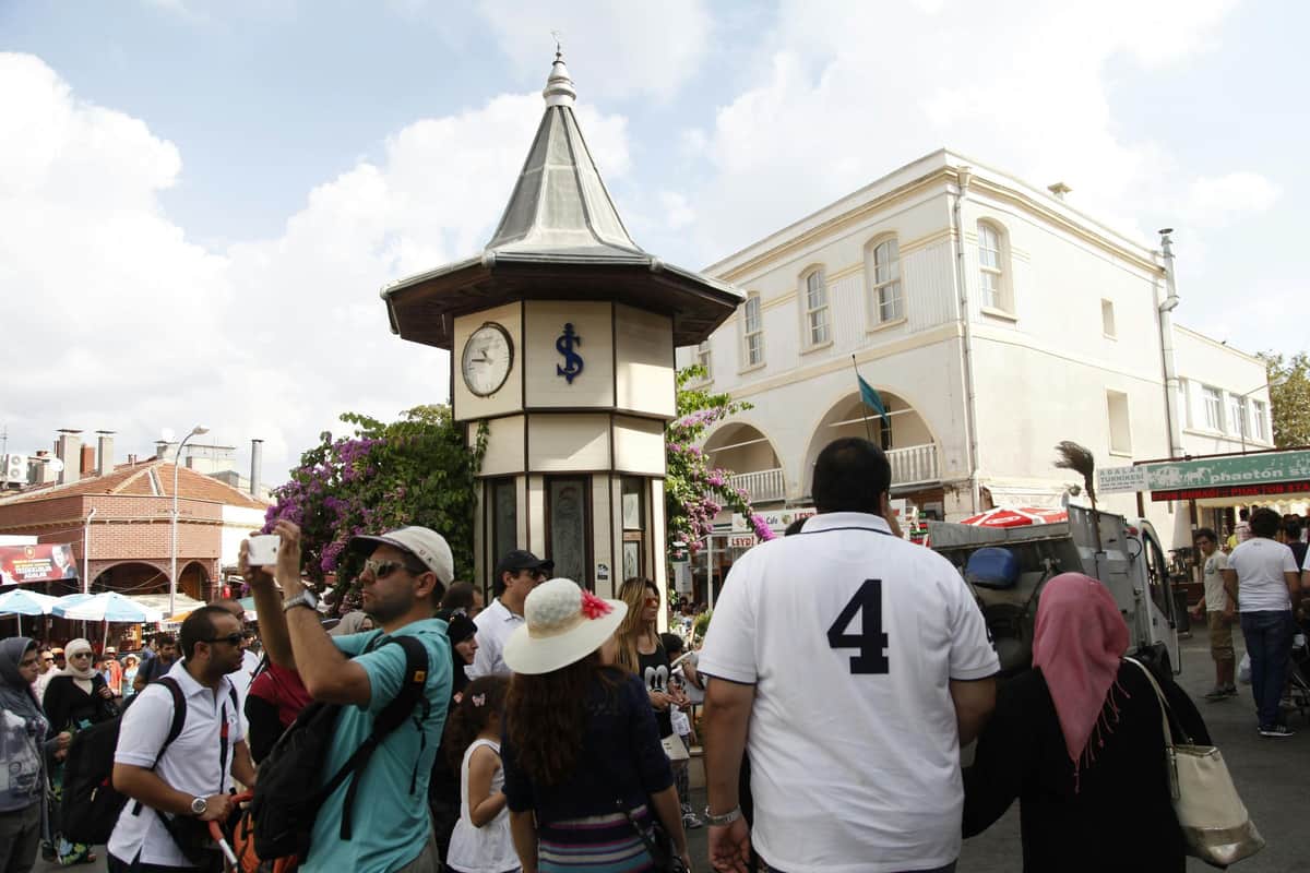 Istanbul: Princes' Islands Private Boat Tour + Optional Pickup & Lunch People walking and taking photos near a small clock tower with a blue dollar sign, under a partly cloudy sky.