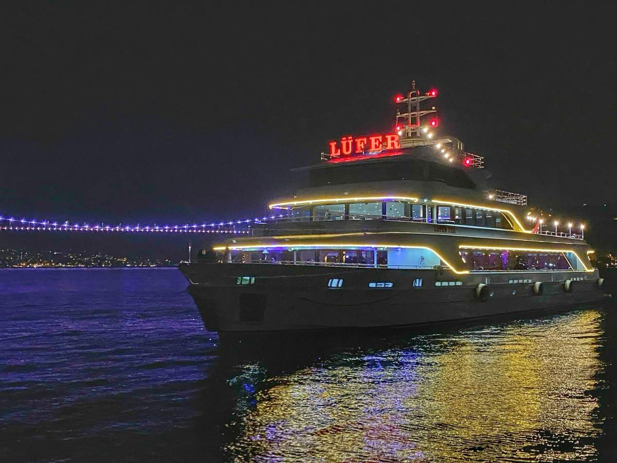A yacht with illuminated lights and the word 'Lüfer' on top is moored near a lit bridge at night.
