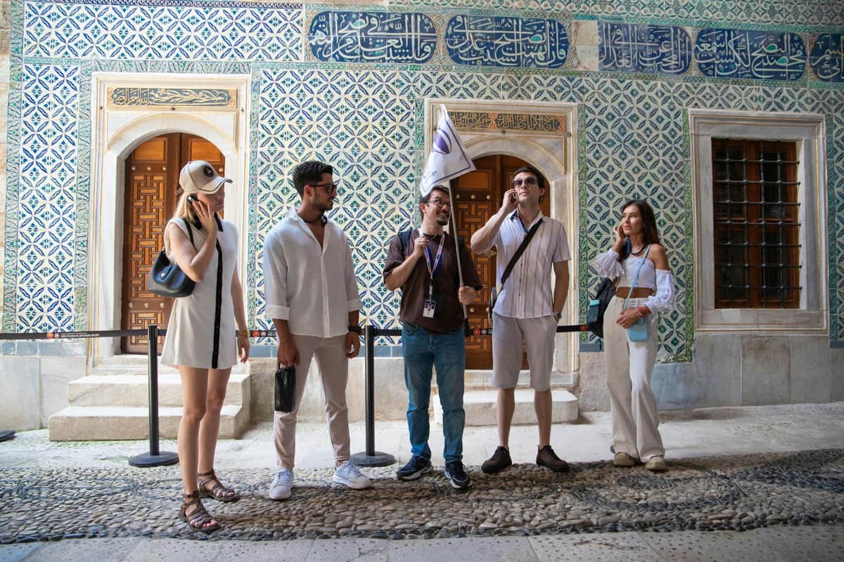 Topkapi Palace: Skip The Line Ticket + Audio Guide + Bosphorus Cruise Four people stand in front of a patterned wall with Arabic script. One person holds a flag, possibly leading a tour.