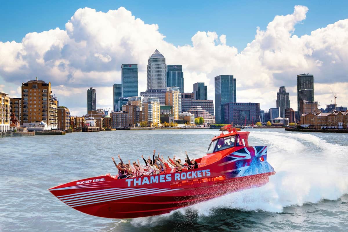 Een rode speedboot met het opschrift „Thames Rockets”, met passagiers in veiligheidsuitrusting, tegen de achtergrond van een stadsgezicht en een gedeeltelijk bewolkte lucht.