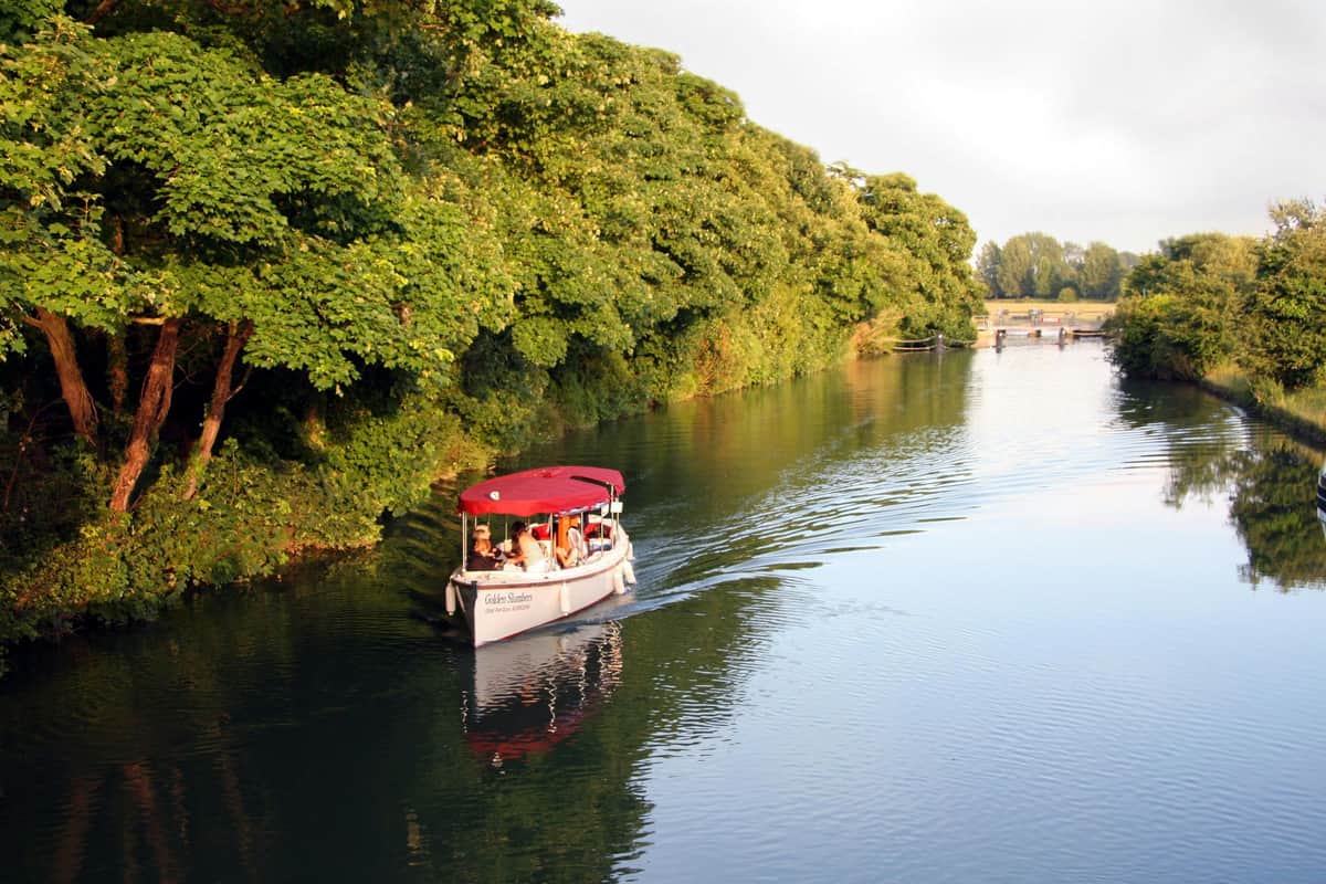Croisière avec thé de l'après-midi à Oxford Un petit bateau avec une verrière rouge navigue sur une rivière à côté d'arbres verdoyants. Les passagers sont assis à l'intérieur.