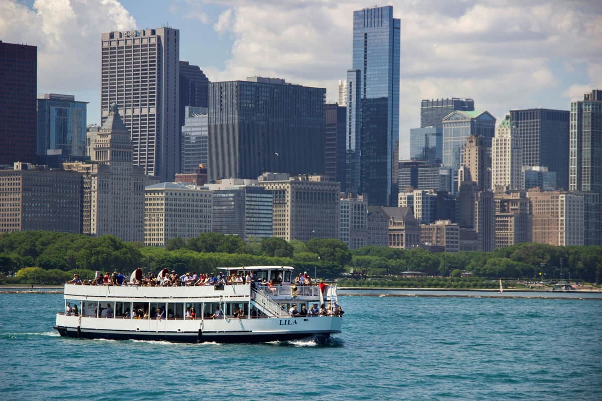 Chicago : visite architecturale de 90 minutes sur le lac et la rivière Un bateau avec des passagers sur une rivière, avec une ligne d'horizon de la ville composée de grands bâtiments et d'arbres en arrière-plan, sous un ciel partiellement nuageux.