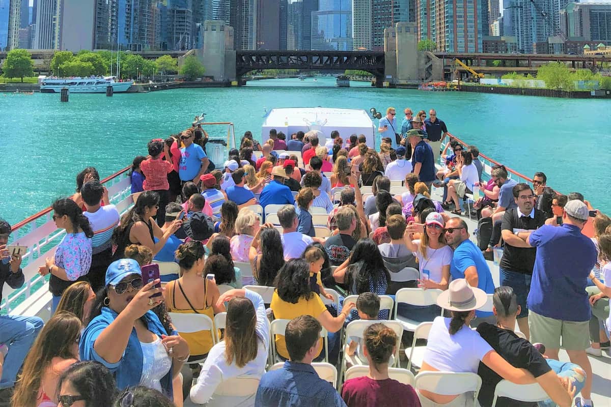 Een groepje mensen zit op een boot en geniet van een zonnige dag, met op de achtergrond de skyline van de stad en een brug over een rivier.