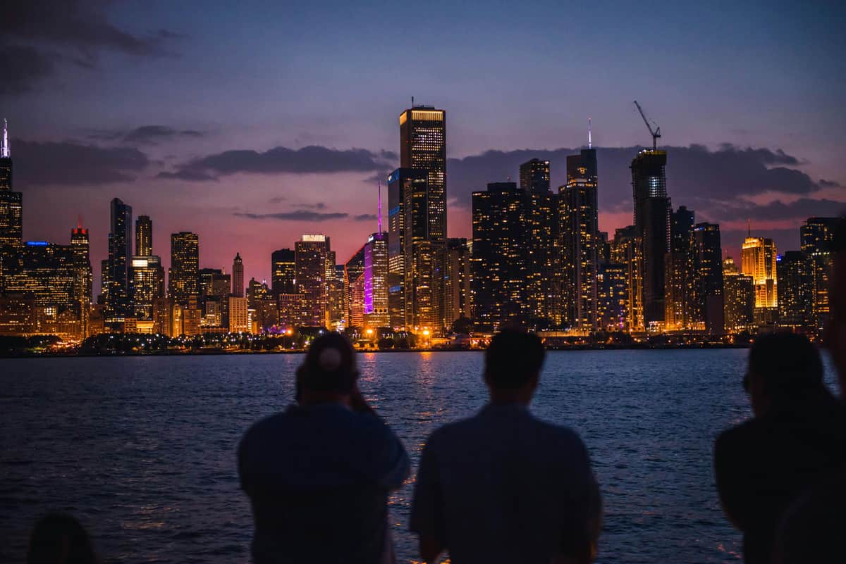 Lac Michigan et rivière Chicago : croisière touristique nocturne de 1,5 heure Deux personnes face à une ville illuminée au crépuscule, avec des bâtiments reflétant la lumière sur une étendue d'eau calme.