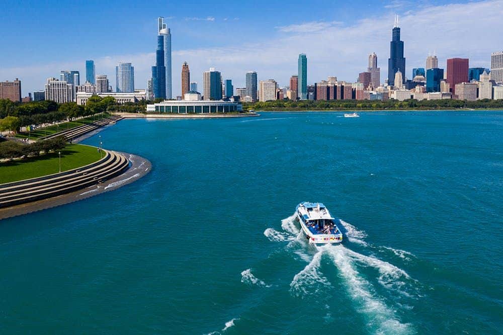 Lac Michigan et rivière Chicago : croisière touristique de 1,5 heure Un bateau navigue sur des eaux bleues avec, en arrière-plan, la silhouette d'une ville, y compris de grands gratte-ciel, sous un ciel bleu clair.
