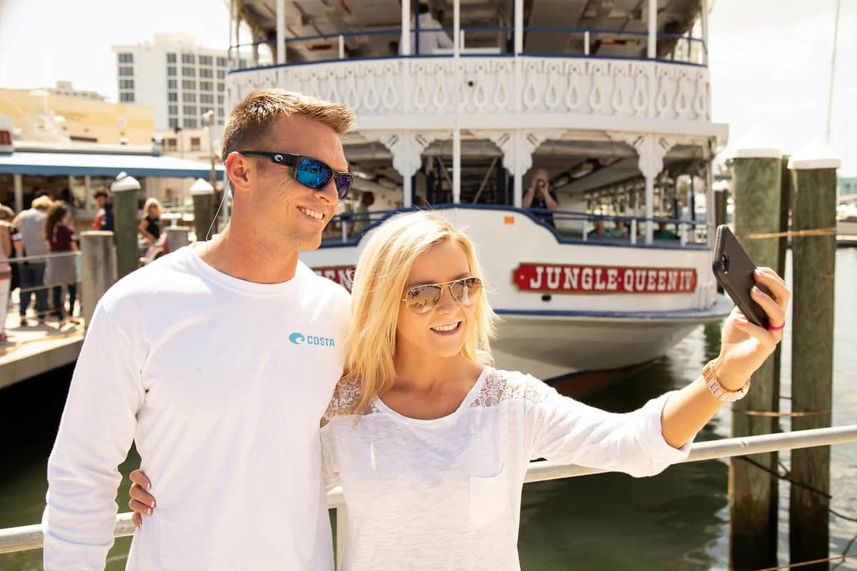 Fort Lauderdale: Jungle Queen Sightseeing Cruise Two people wearing sunglasses smile for a photo in front of a boat named "Jungle Queen" docked at a marina.