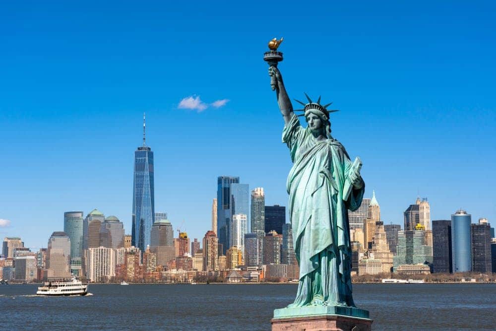 Het Vrijheidsbeeld met de wolkenkrabbers van New York City, waaronder het One World Trade Center, op de achtergrond onder een strakblauwe hemel.