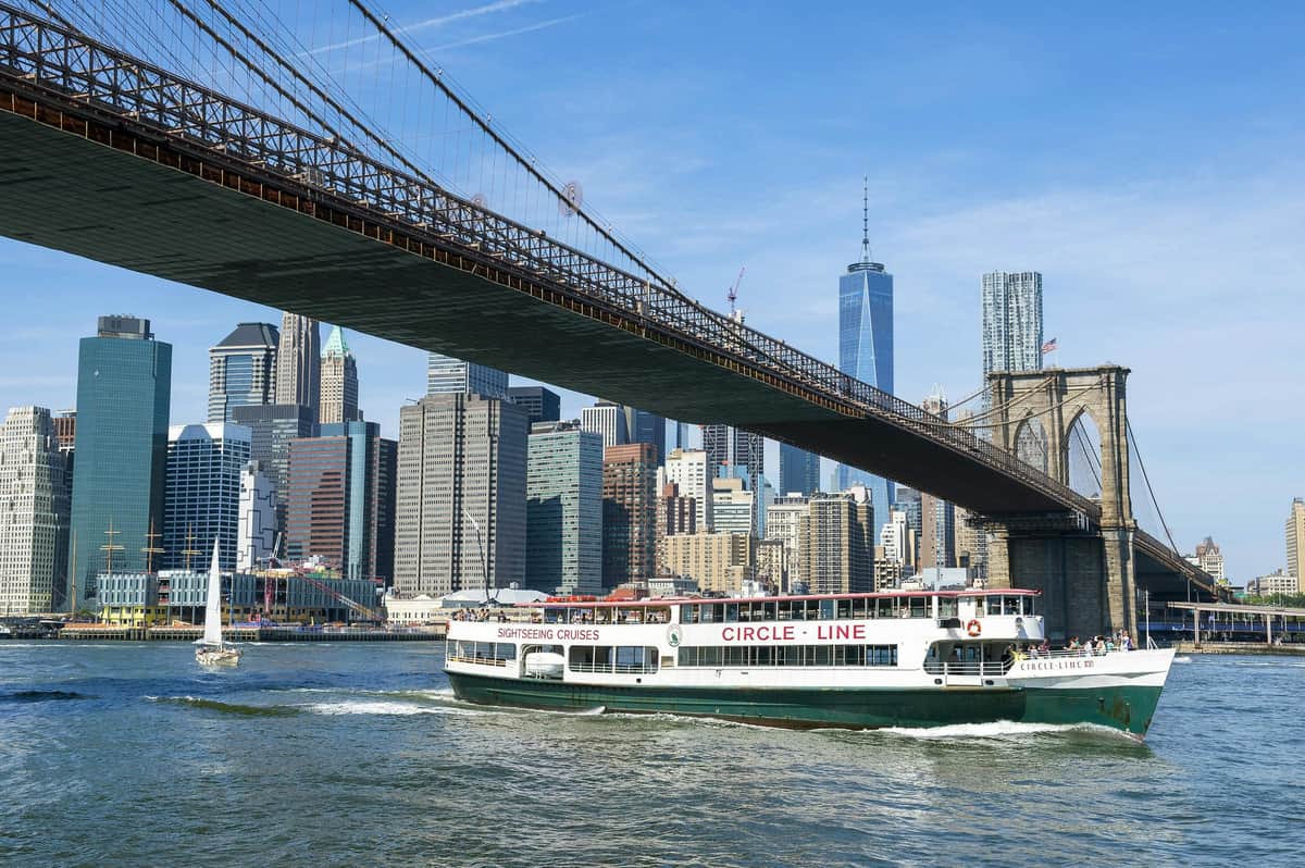 Een rondvaartboot met het label "Circle Line" vaart onder de Brooklyn Bridge door met de skyline van New York City op de achtergrond.