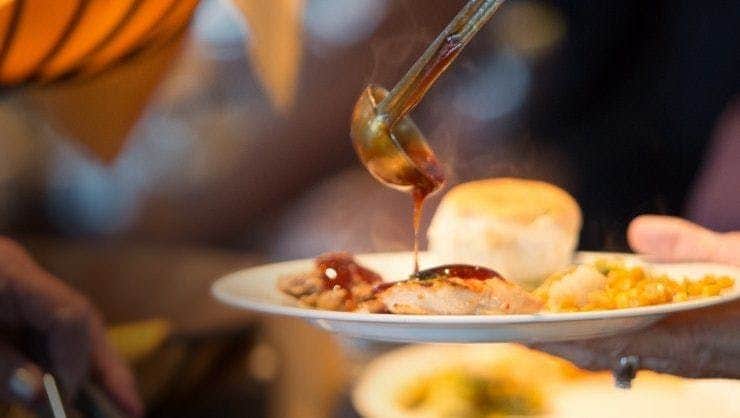 Captain's Lunch Cruise A ladle pours sauce over a meal on a plate, which includes a piece of meat and a bread roll. Steam rises from the food.