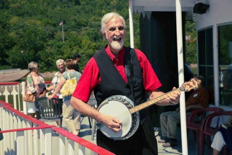 Historic Cincinnati Sightseeing Cruise A man with white hair and beard plays a banjo on a sunny outdoor patio with people talking in the background.