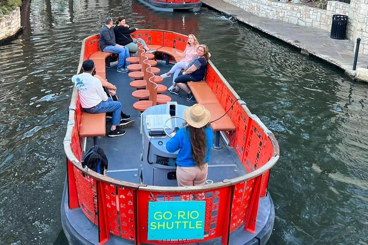 San Antonio: 1- of 3-daagse River Shuttle-pas Een rivierpendelboot met passagiers die aan de zijkanten zitten en een gids die vooraan aan het roer staat, met het opschrift ‘GO RIO SHUTTLE’.