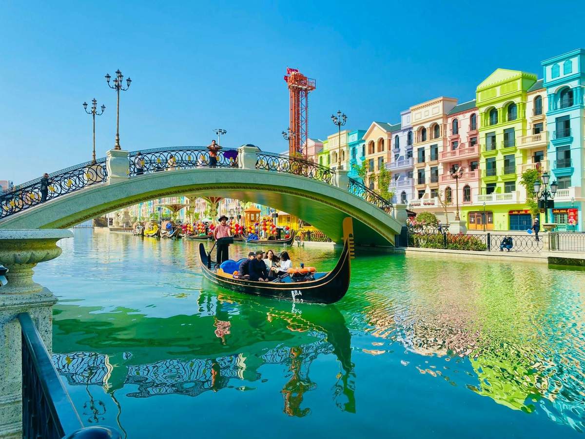 Una gondola con dei passeggeri su un canale, circondata da edifici colorati e da un ponte decorativo sotto un cielo azzurro e limpido.
