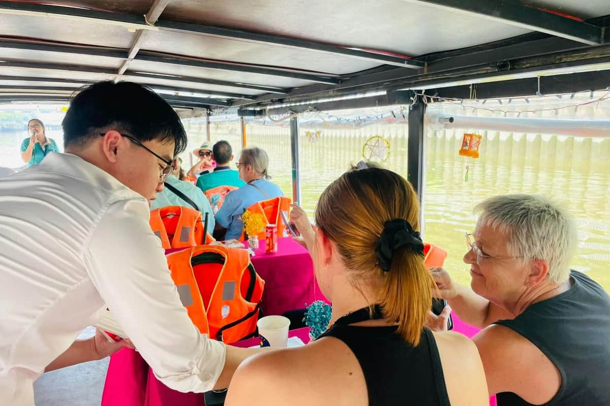 Ho Chi Minh Ville : Croisière sur le canal Nhieu Loc-Thi Nghe + boisson Personnes assises à des tables sur un bateau fluvial, certaines portant des gilets de sauvetage orange. Un serveur en chemise blanche s'occupe des passagers.