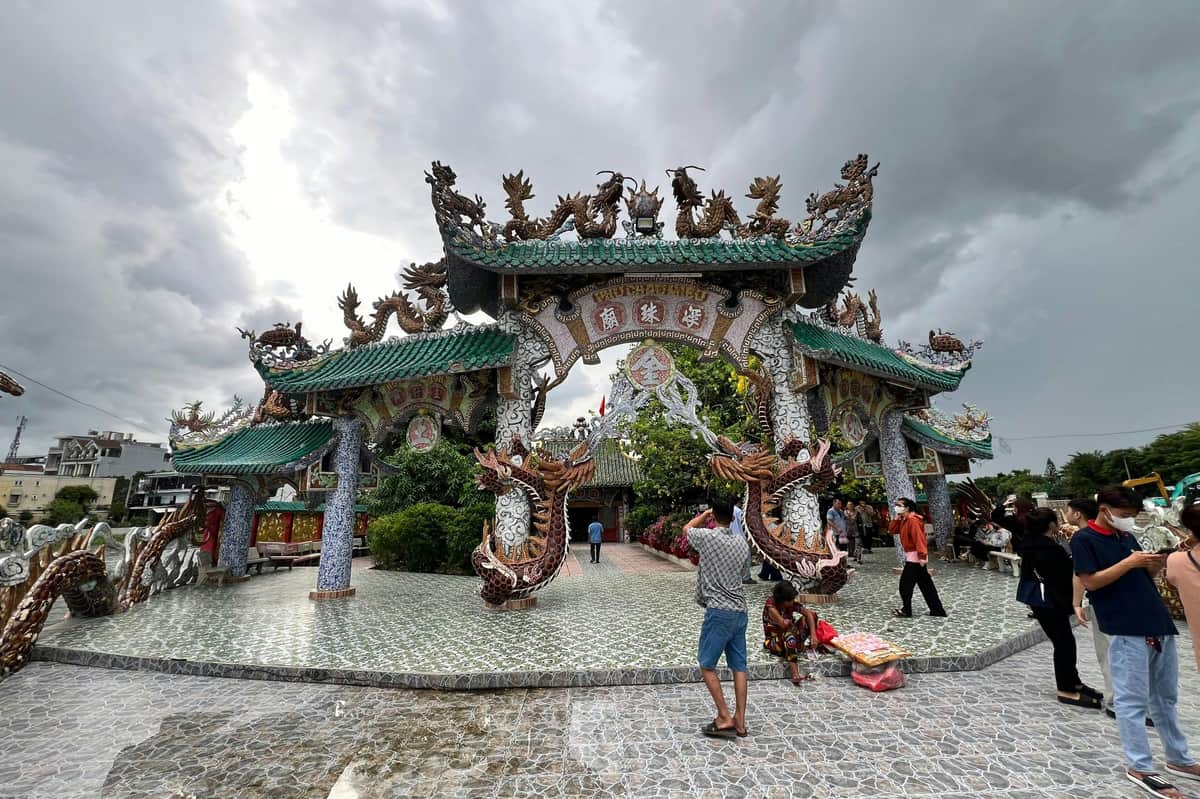 Temple flottant de Phu Chau et village touristique de Binh Quoi : Excursion guidée en hors-bord Une porte ornée de décorations de dragons dans une zone carrelée, avec des gens qui marchent et prennent des photos, sous un ciel nuageux.