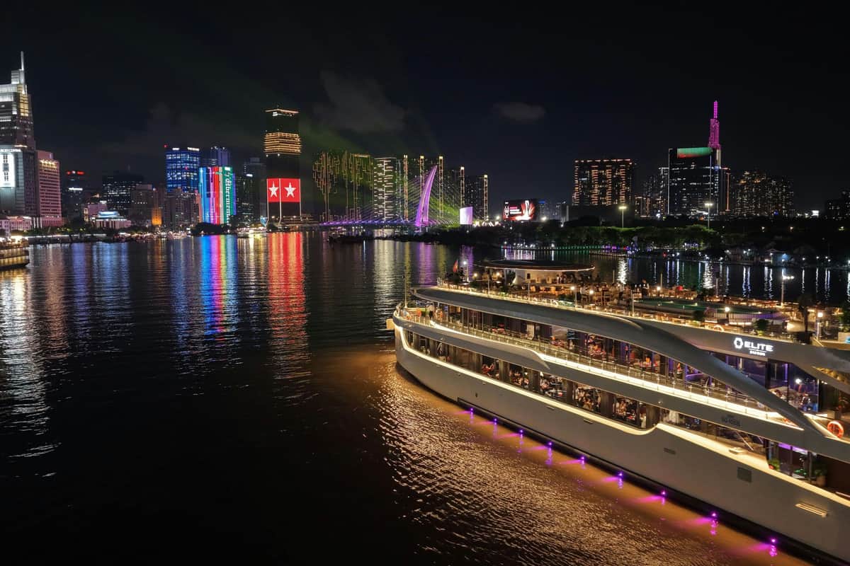 Saigon River: Dinner Cruise + Drinks A brightly lit cruise ship docked on a river at night with a cityscape of illuminated skyscrapers reflecting in the water.