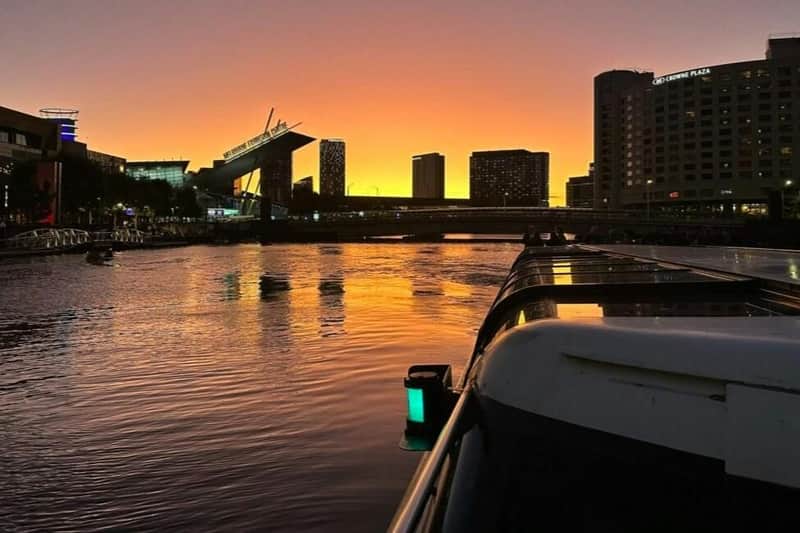 Zonsondergang over een stadslandschap met moderne gebouwen en een rivier op de voorgrond, inclusief een boot die aan de zijkant is aangemeerd.