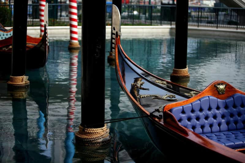 The Venetian Macao: Gondola Ride + Free Legend Heros Park 5 Game Points A gondola with a polished wooden finish and blue seats docked near striped poles on calm water.