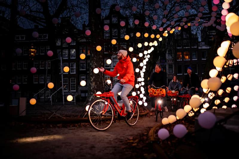 Mensen fietsen door een park versierd met gloeiende bolvormige lampjes tijdens de schemering, met gebouwen op de achtergrond.