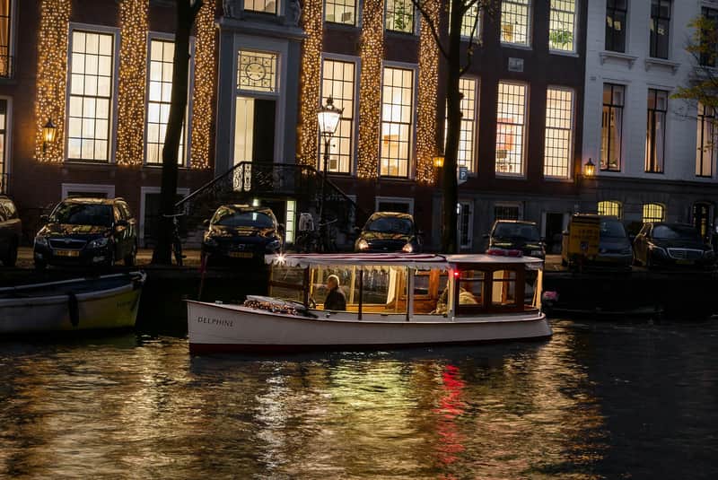 Een boot met de naam "Delphine" op een kanaal met verlichte gebouwen erachter en geparkeerde auto's langs de waterkant in de schemering.