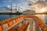 A boat with orange cushions and flowers, a table with drinks, a historic ship, and a large building by the water.