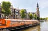 An orange "Lovers Canal Cruises" boat on a canal, with historic buildings and a church tower in the background.