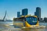 A yellow amphibious bus travels on water in front of a cityscape with modern skyscrapers under a clear blue sky.